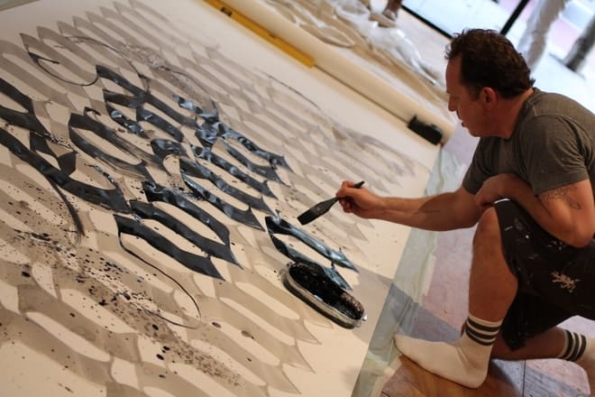 Niels Shoe Meulman painting calligraffiti on a large paper laying on the ground with black paint