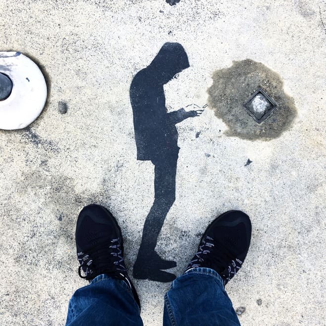 Stencil graffiti on the ground of a figure standing and looking at his phone